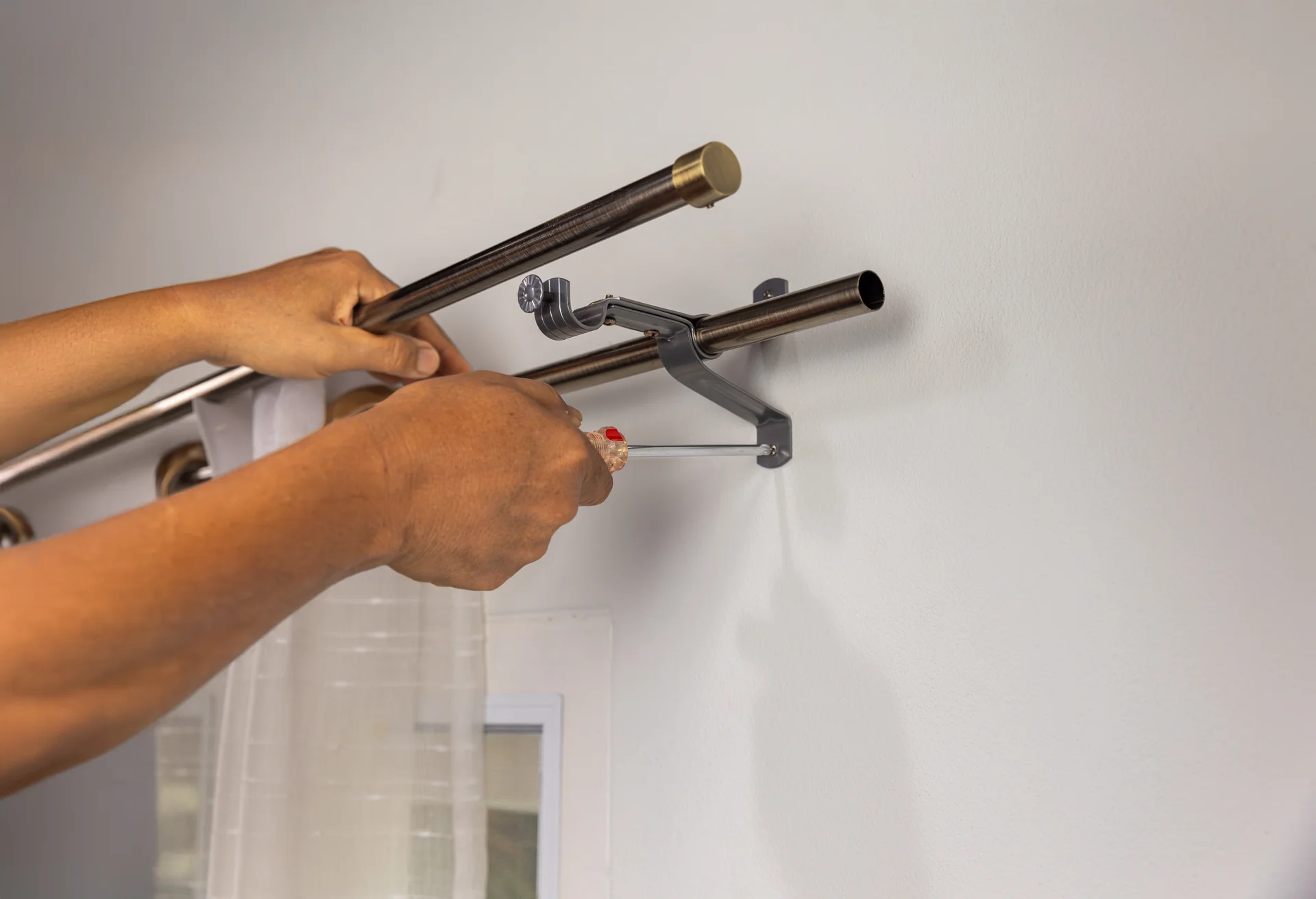 Hands using a screwdriver to secure a curtain rod bracket to a wall mount during curtain rod installation